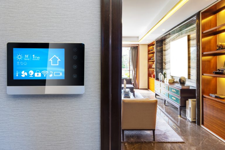 A touchscreen thermostat mounted on the wall and a modern living room on the background, Should You Ventilate Your Home in Winter?
