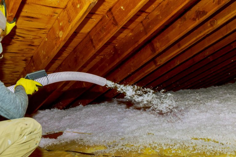 An insulation specialist pouring in blown in insulation in the flooring of an attic, Is Blown-In Insulation Cheaper Than Rolls?