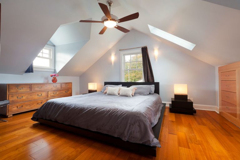 Interior of a rustic themed mansard bedroom with polish wooden flooring and white wall and ceiling, Do Ceiling Fans Help AC?