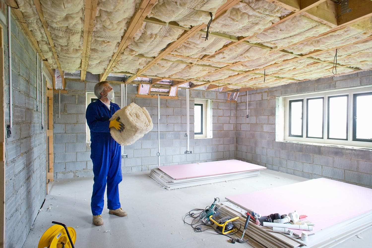 A man in coveralls holding ceiling insulation and looking up in an under construction house
