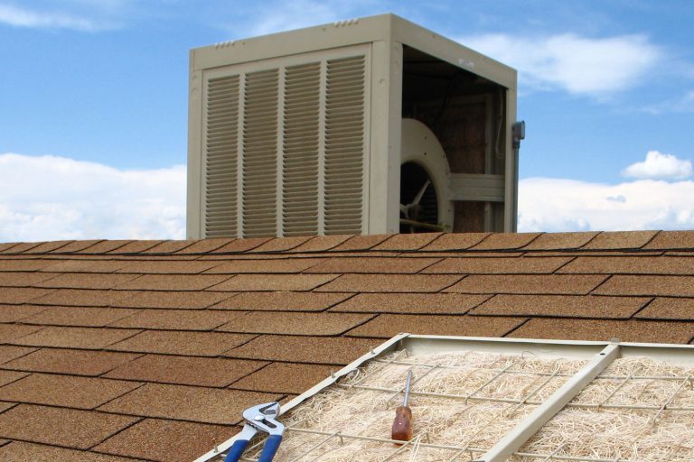 This color photo shows a swamp cooler on a reddish-orange shingled roof. One panel of the swamp cooler has been removed and placed in the foreground to show the pad or filter made of aspen wood, Swamp Cooler Spraying Water - What To Do?