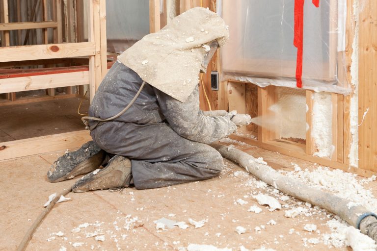 A worker spraying foam insulation on the wall of a house under construction, How Long Does Spray Foam Insulation Smell?