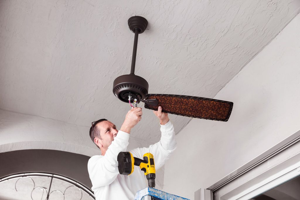 electricians hanging a ceiling fan. The man is holding wires in his hand. He is wearing a long sleeved white shirt and is fixing the fan. He has a drill sitting on a ladder