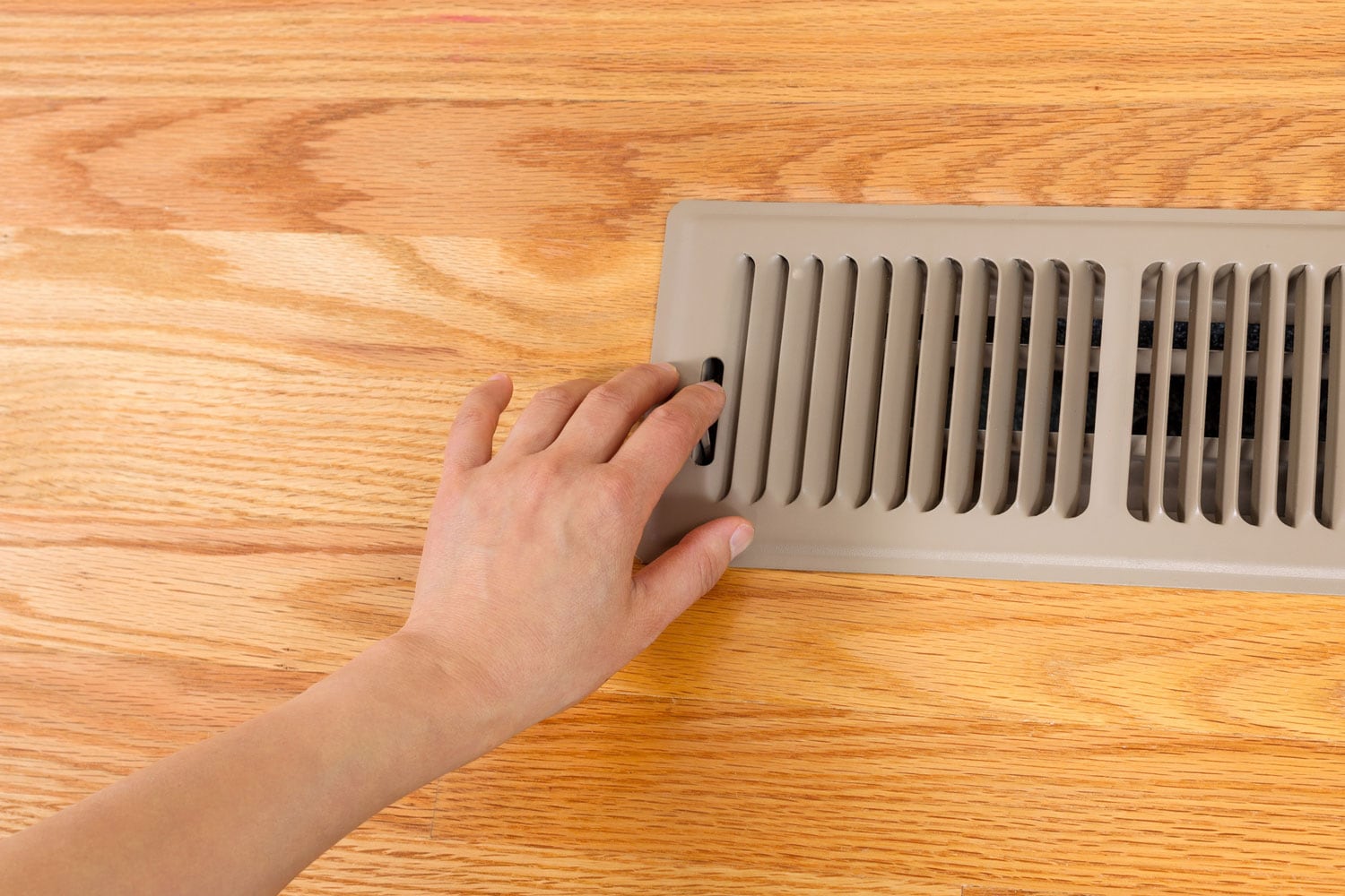 Horizontal photo of female hand opening up heater floor vent with Red Oak Floors in background