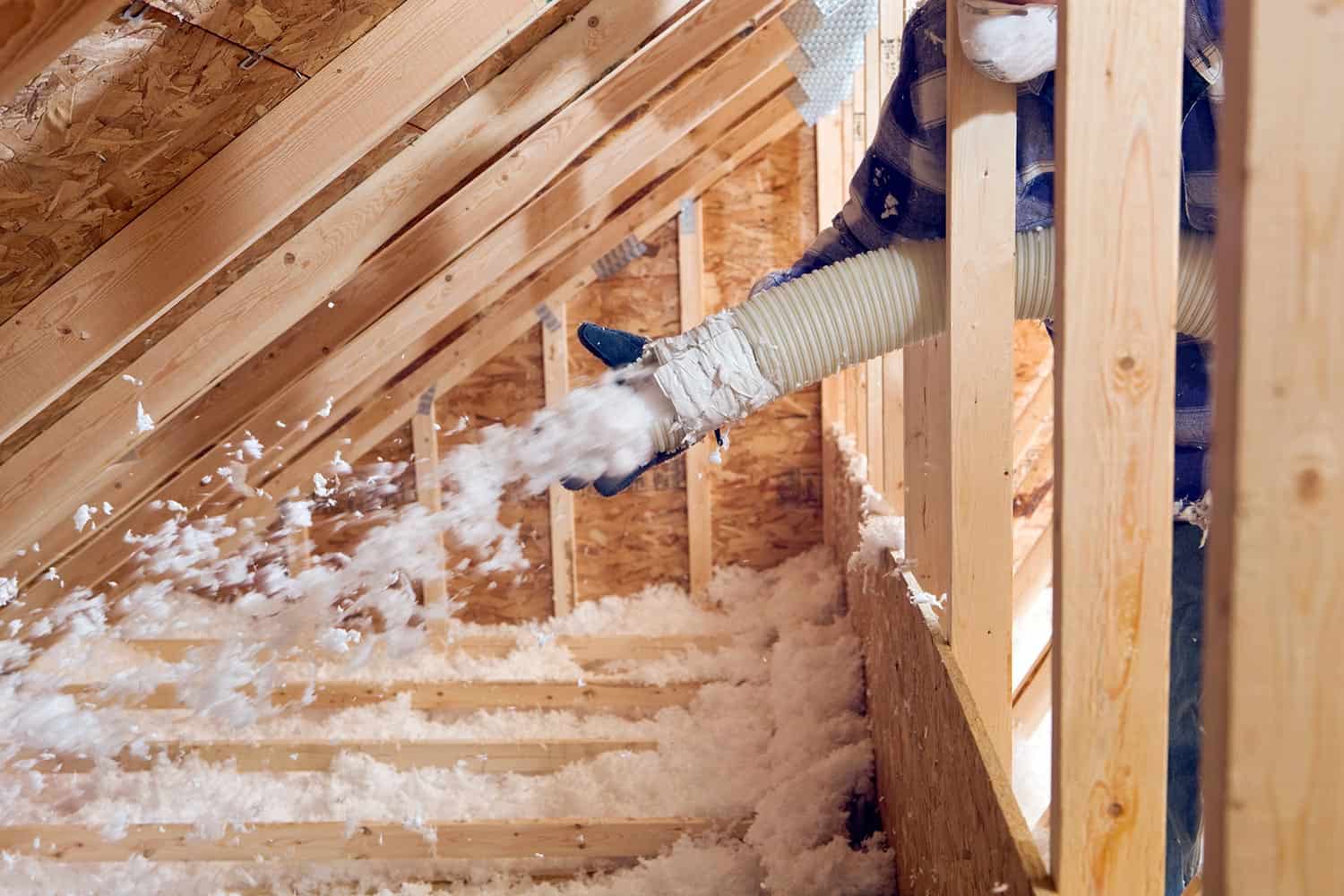 Worker spraying blown fiberglass insulation between attic trusses