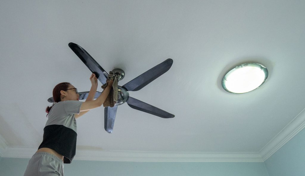 An Asian woman cleaning ceiling fan at home.