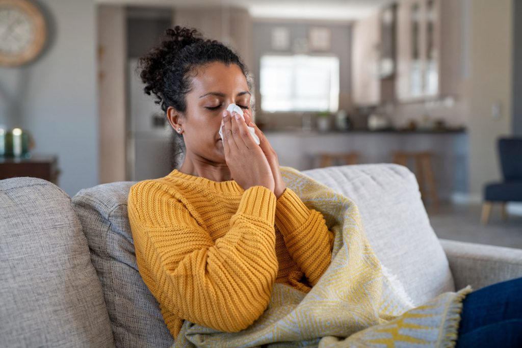 Sick african woman wrapped in blanket sitting on sofa blowing her nose at home