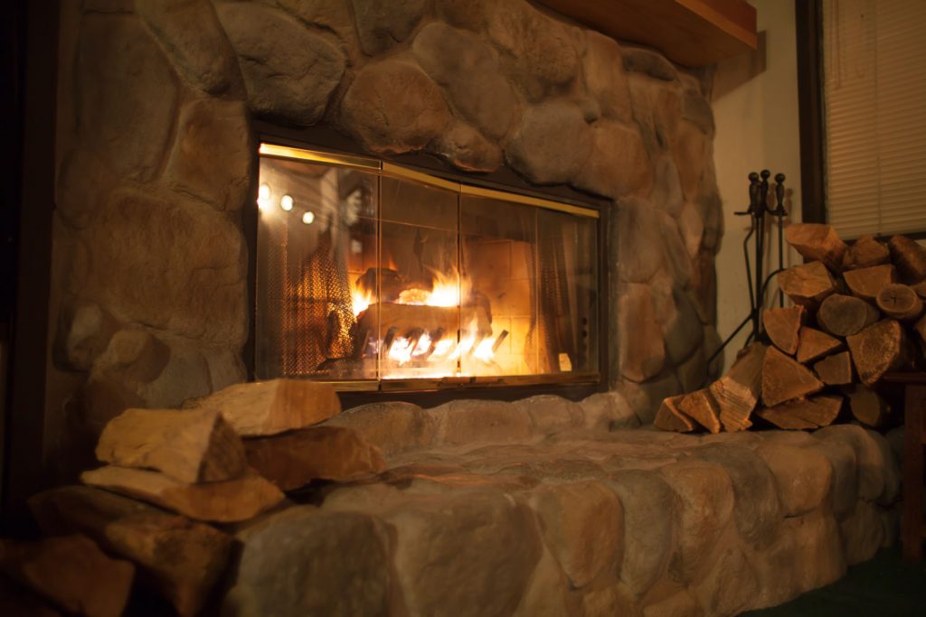 A stone decorated fireplace with firewood on the side and a glass covered fireplace