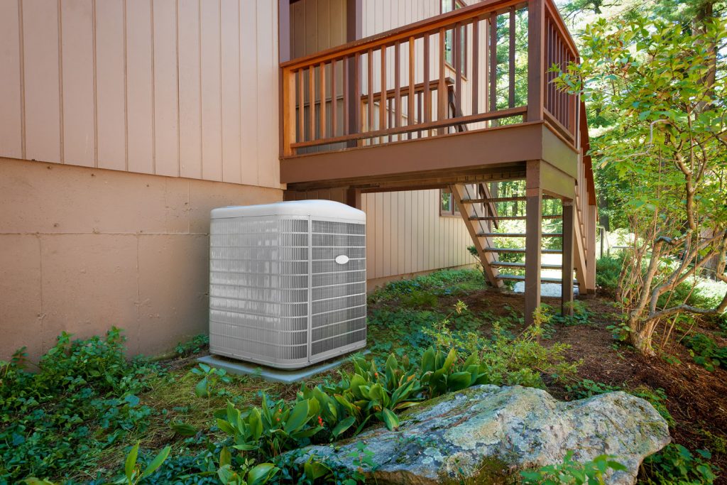 A white heat pump unit placed outside the house under the entryway stairs