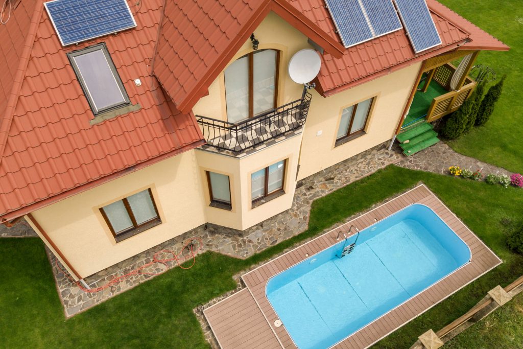 Aerial view of a new autonomous house with solar panels and water heating radiators on the roof and green yard with blue swimming pool.