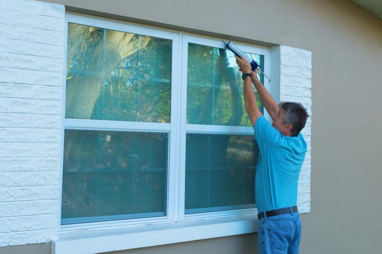 Homeowner caulking window with a caulk gun, How To Insulate Very Large Windows [For Summer And Winter]