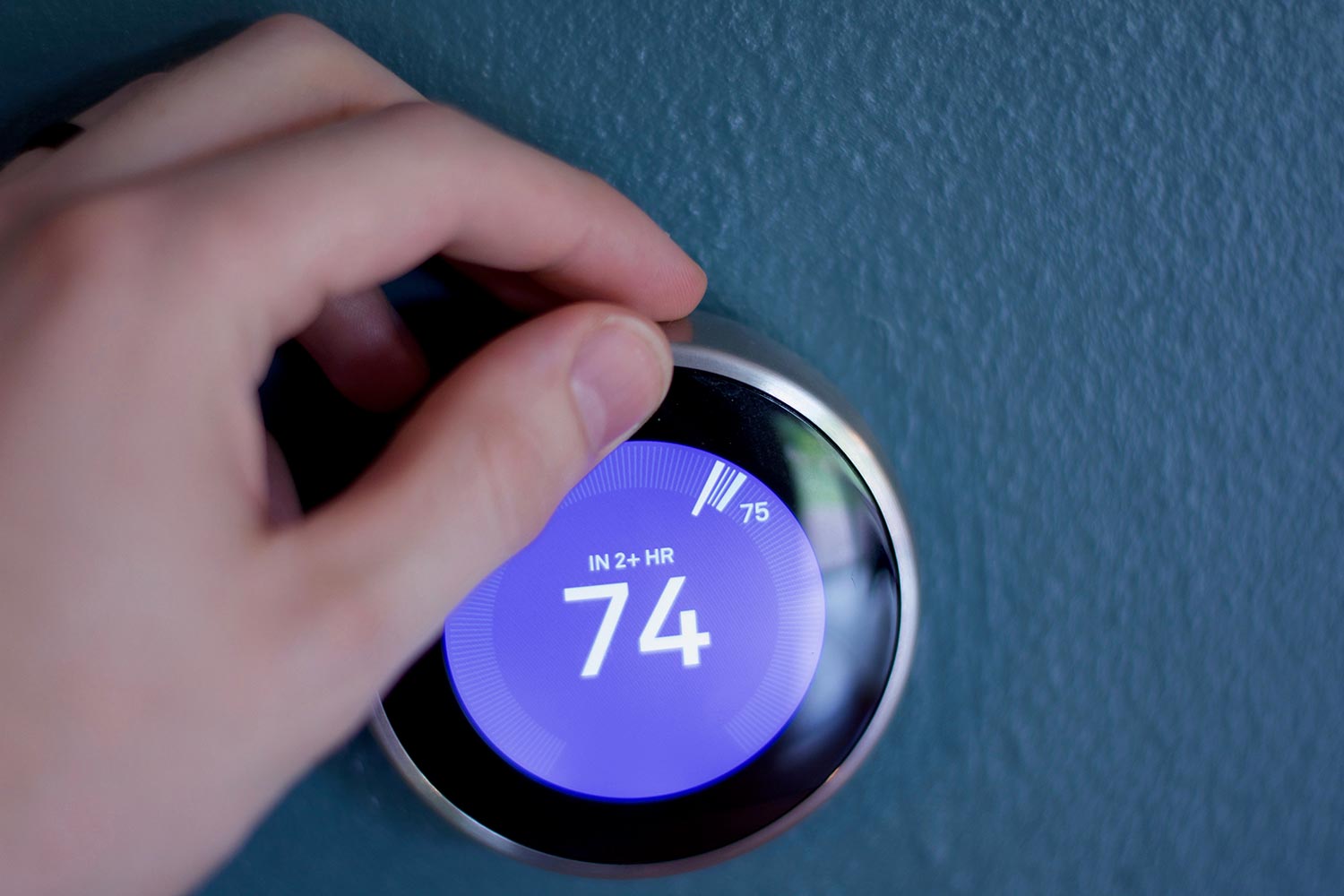 Smart thermostat being adjusted by male hand against a blue wall