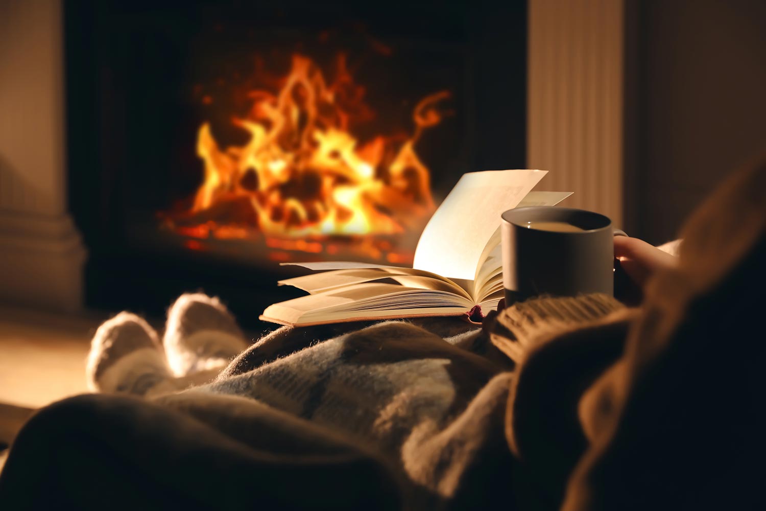 Woman with cup of drink and book near fireplace at home