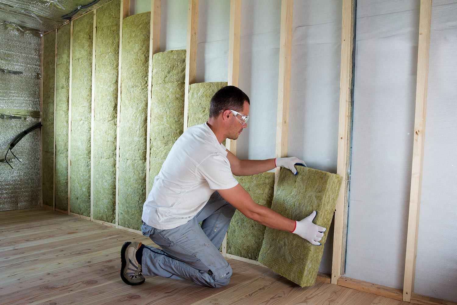 Worker in protective goggles and respirator insulating rock wool insulation in wooden frame for future house walls for cold barrier