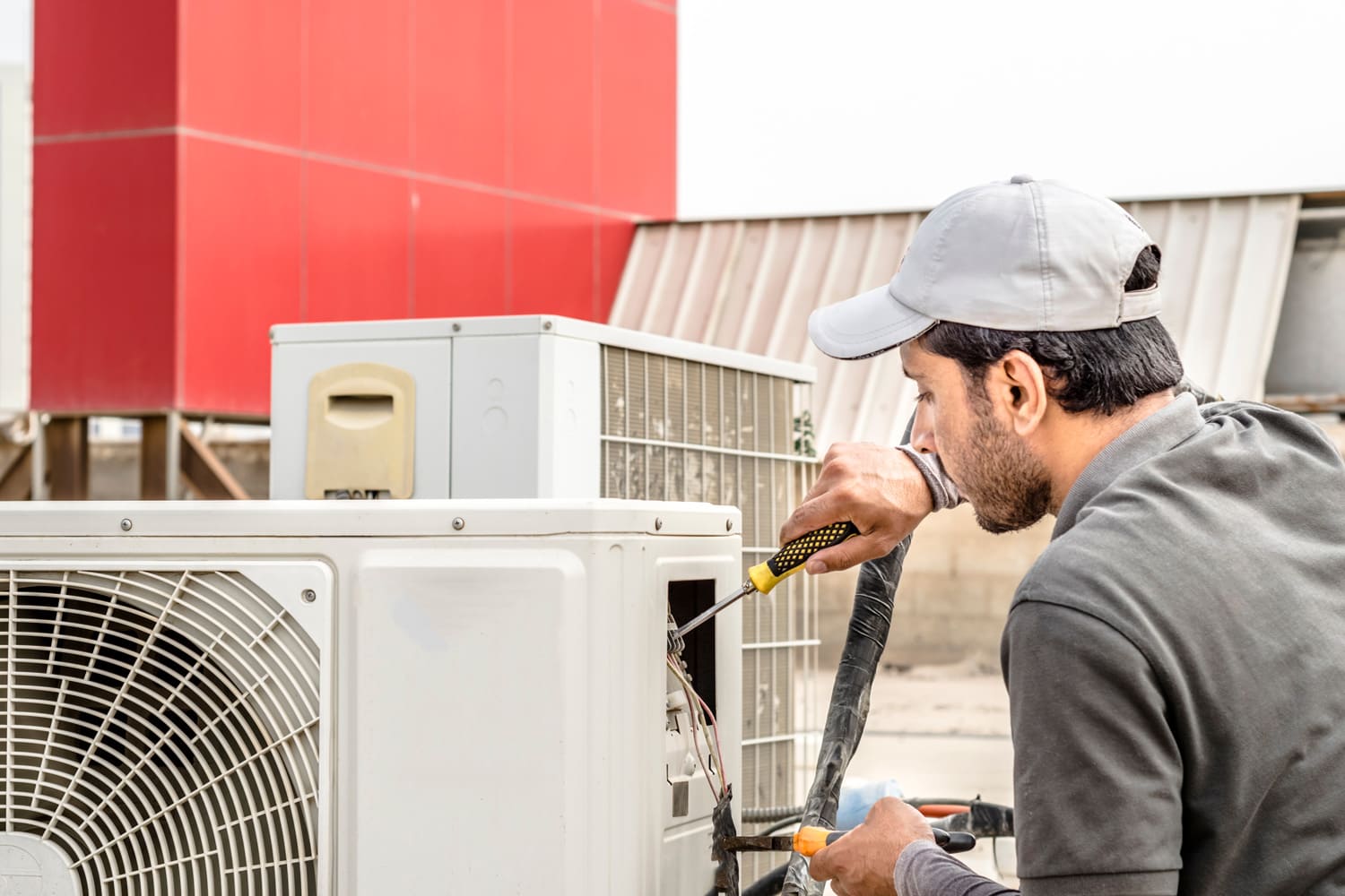 a professional electrician man is fixing a heavy duty unit of central air conditioning system by his tools on the roof top and wearing grey color of uniform and white cap