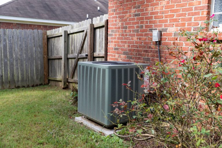 Air conditioner condenser unit sitting next to brick home with fence, Amana Air Conditioner Won't Turn On - What To Do?