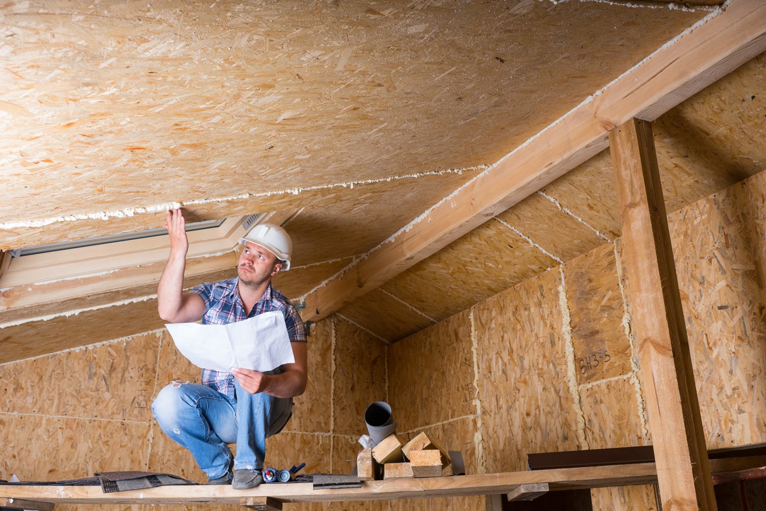 Male Construction Worker Builder Wearing White Hard Hat Crouching on Elevated Scaffolding and Reading Plans near Ceiling of Unfinished Home with Exposed Plywood Particle Board