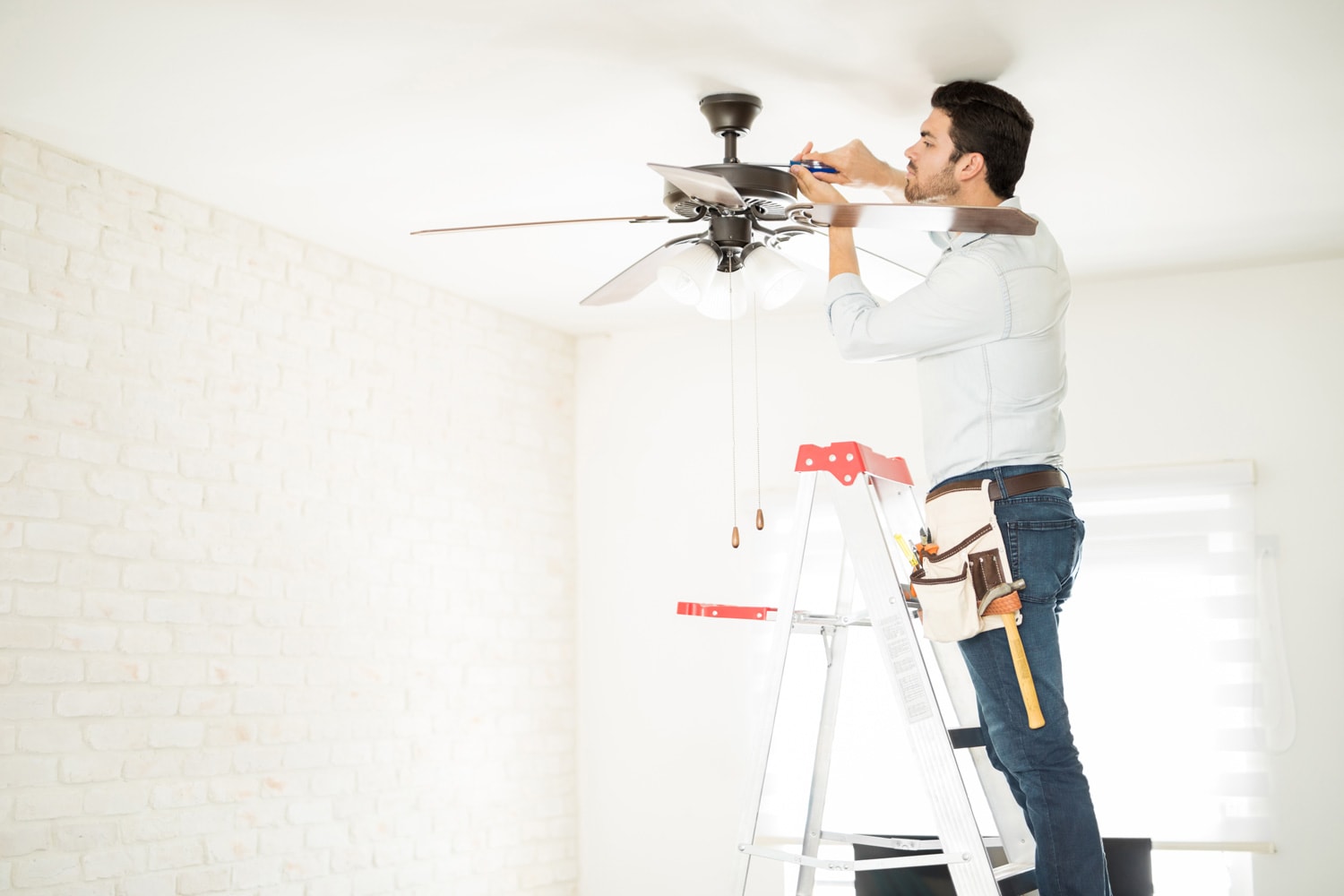 Profile view of a male electrician stepping on a ladder and installing a ceiling fan in a house