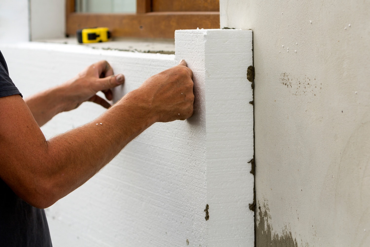 Worker hands installing white rigid polyurethane foam sheet on plastered brick wall.