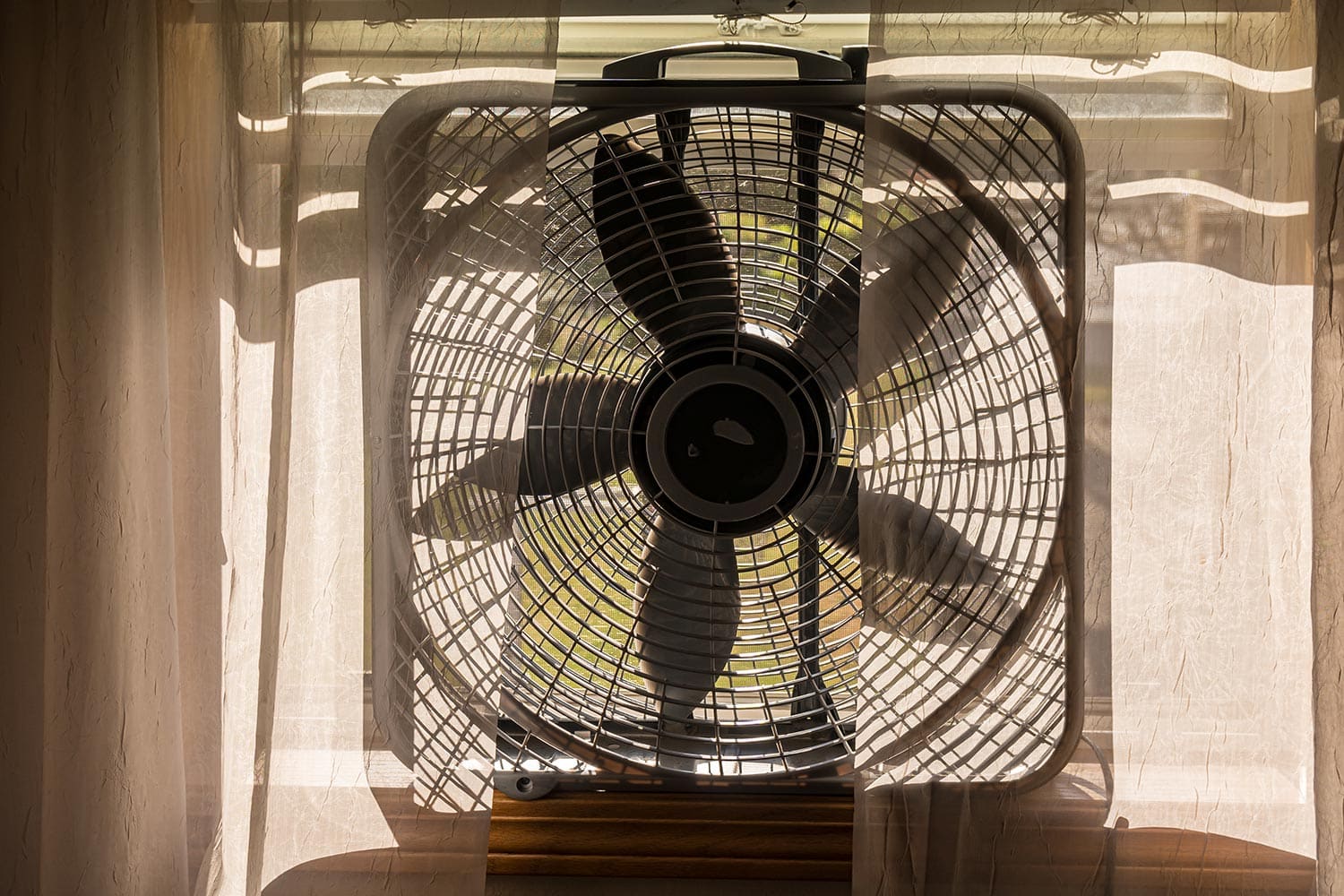 Electric box fan sitting on a window sill with sheer lace curtains