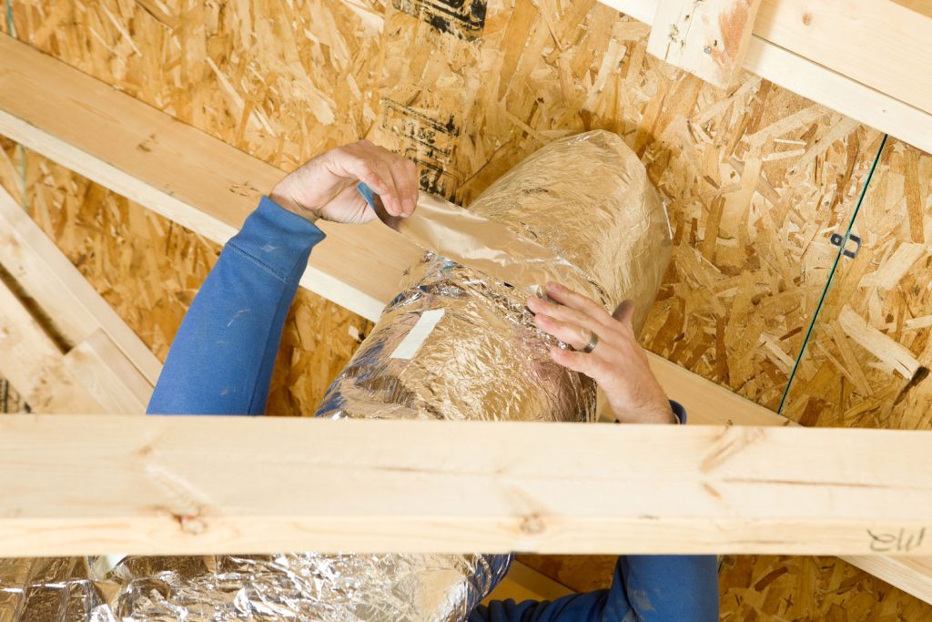 A worker is sealing two sections of house attic vent duct insulation with aluminum foil tape. The foreground is a section of truss and the background is OSB sheathing for the roof.