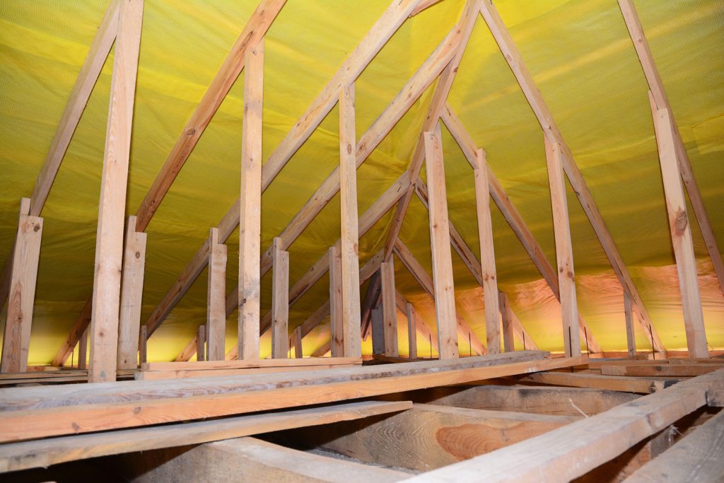 A view on unfinished attic from inside the house with a close-up on wooden ceiling joists, roof beams, rafters, wall studs and vapor barrier film installed under the roof.