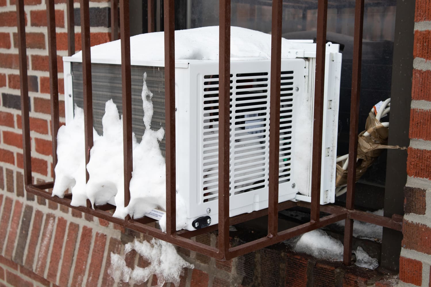 A window air conditioner in a barred cage with snow on a brick urban building in New York City
