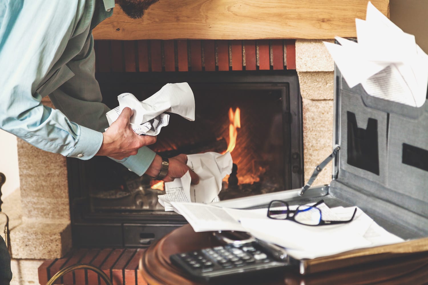 audit documents. Hands of businessman crushes paper documents from an open briefcase before burning fireplace. On top of the papers are fashionable glasses