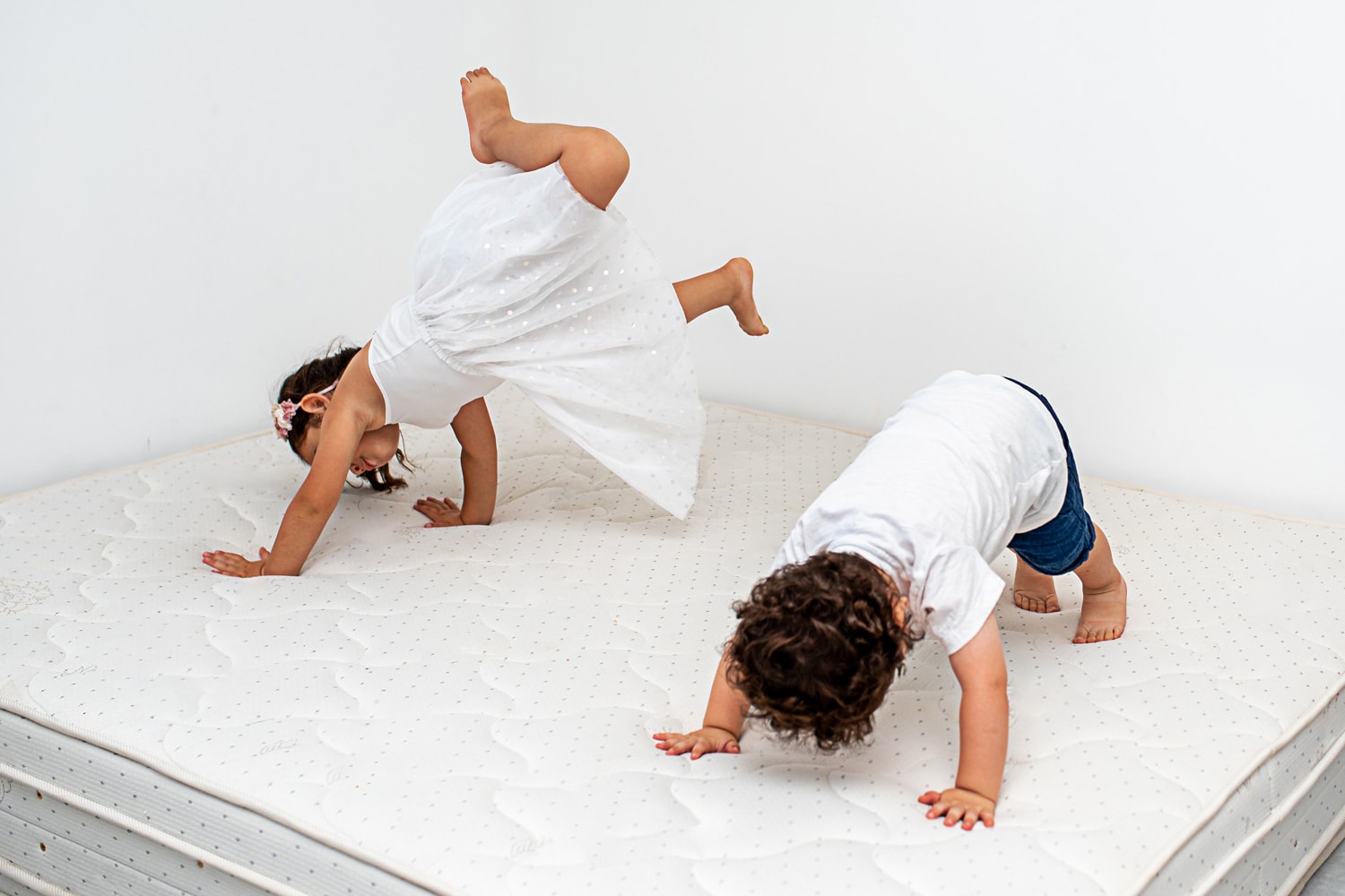 Two crazy cheerful cheery positive preschool siblings having fun, jump on the mattress in a home over white background. Quarantine. Selective focus on girl.