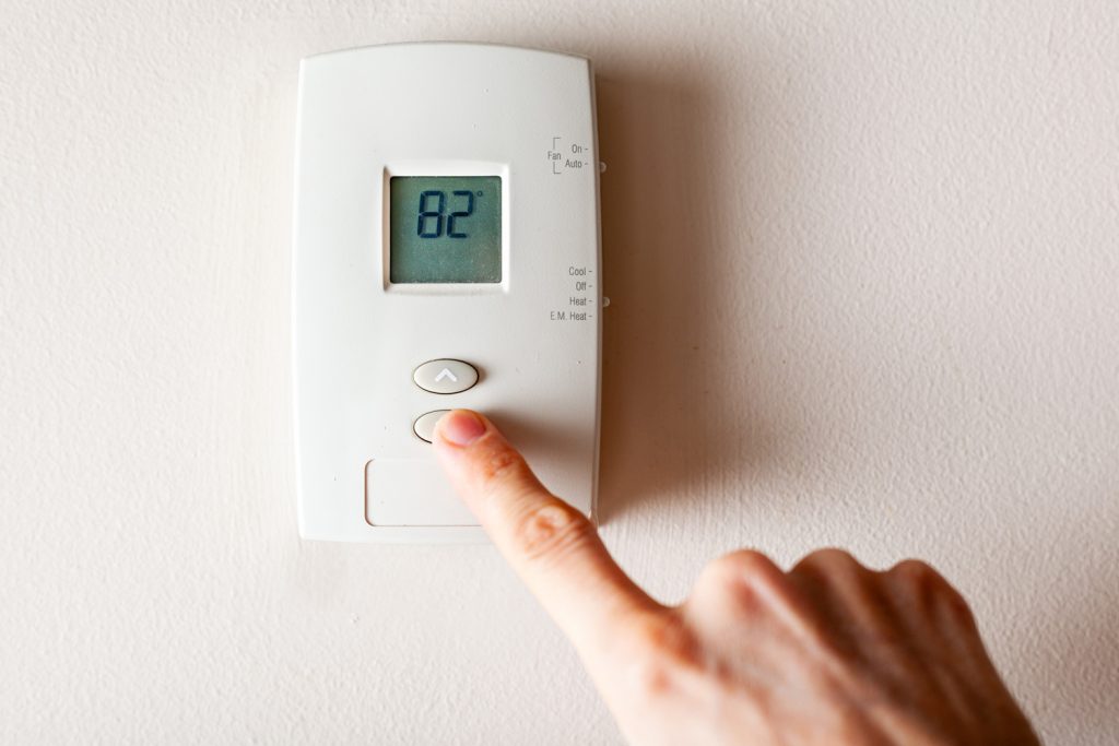 A woman is pressing the down button of a wall attached house thermostat with digital display showing the temperature.