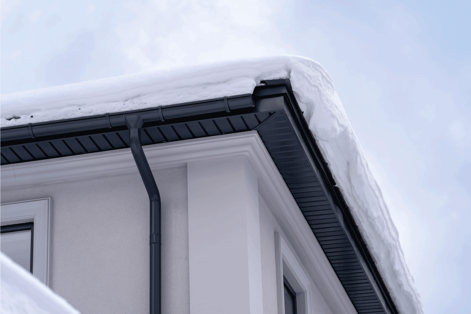 Corner of house with roof made of gray metal tiles and gutter covered with thick layer of snow in winter.