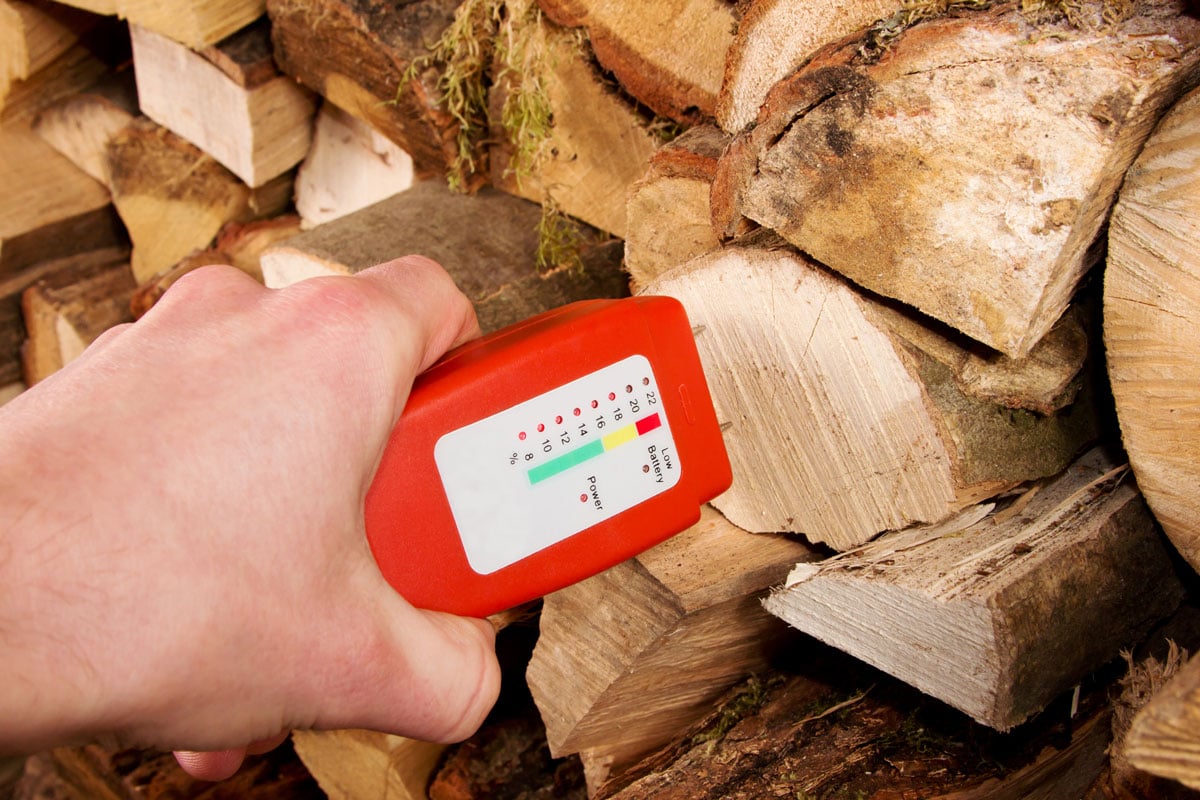 Hand with a wood moisture meter in front of a stack of firewood