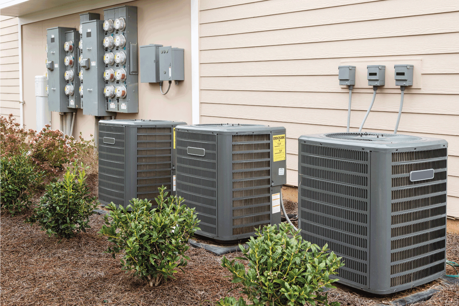 Three air conditioning units with a panel of electricity meters behind, all at the side of a recently-constructed apartment building.