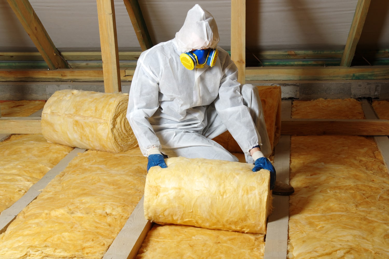 Worker in protective respirator insulating glass wool insulation in wooden construct.