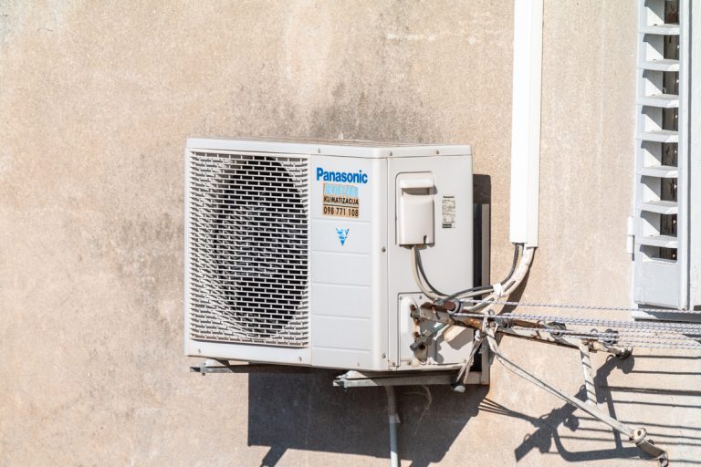 A Panasonic air conditioning inverter mounted on metal brackets behind a building, Panasonic AC Timer Always On - Why And What To Do?