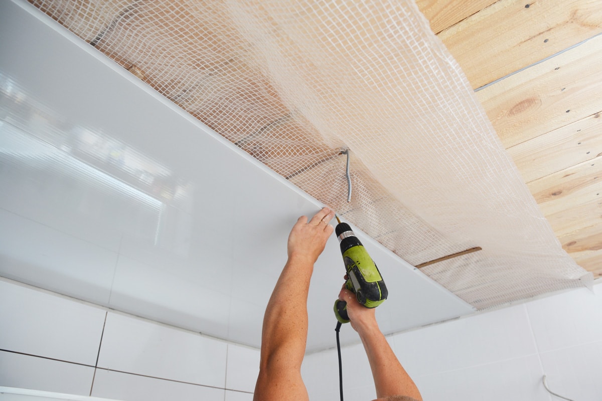 Installing wall and ceiling panels on planked wood ceiling covered with vapor barrier membrane using screwdriver while bathroom renovation, remodel.