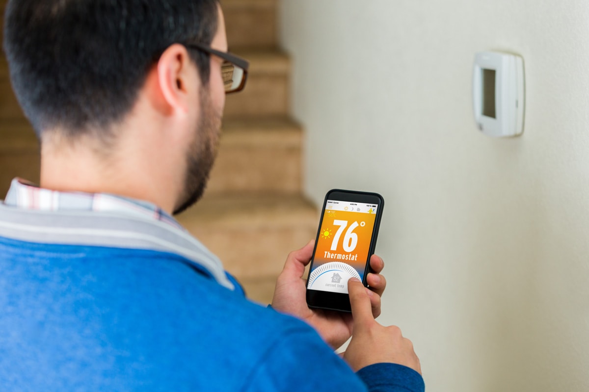 Mid adult male homeowner uses smart phone technology to control his home's temperature remotely. He is standing near the thermostat's control panel.