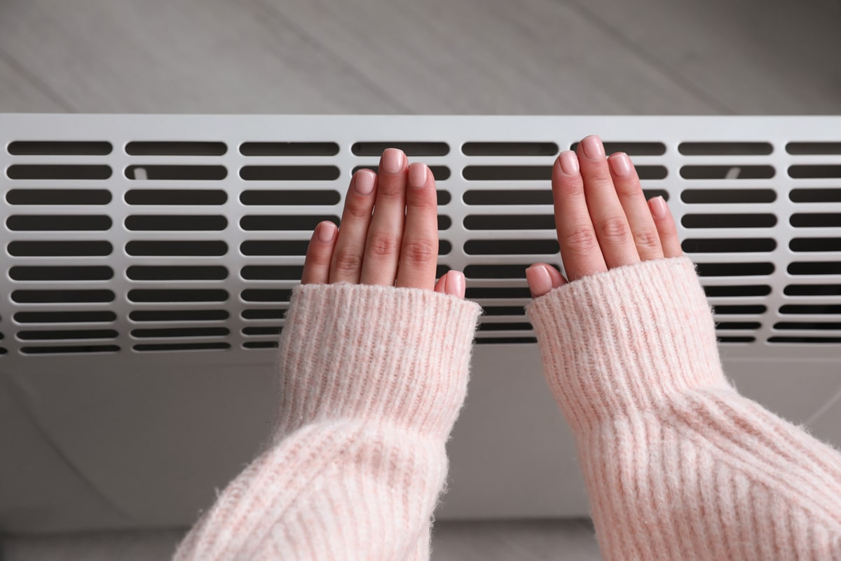 Woman warming hands near electric heater at home, top view