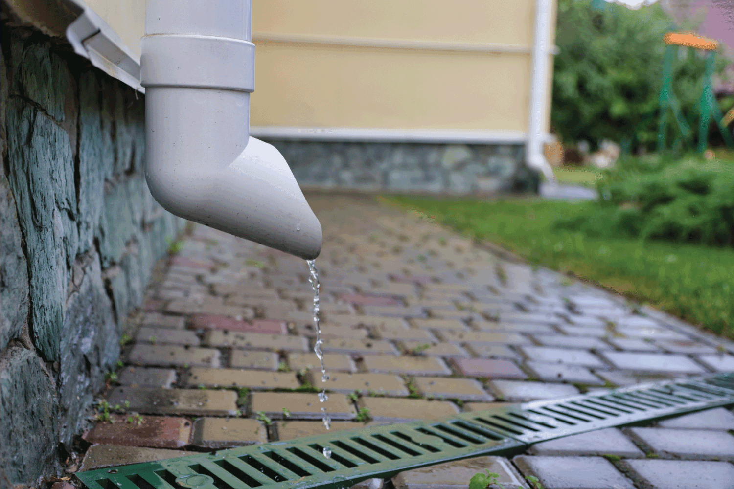 water flows down a white drainpipe of a beige house into a gutter with a grate