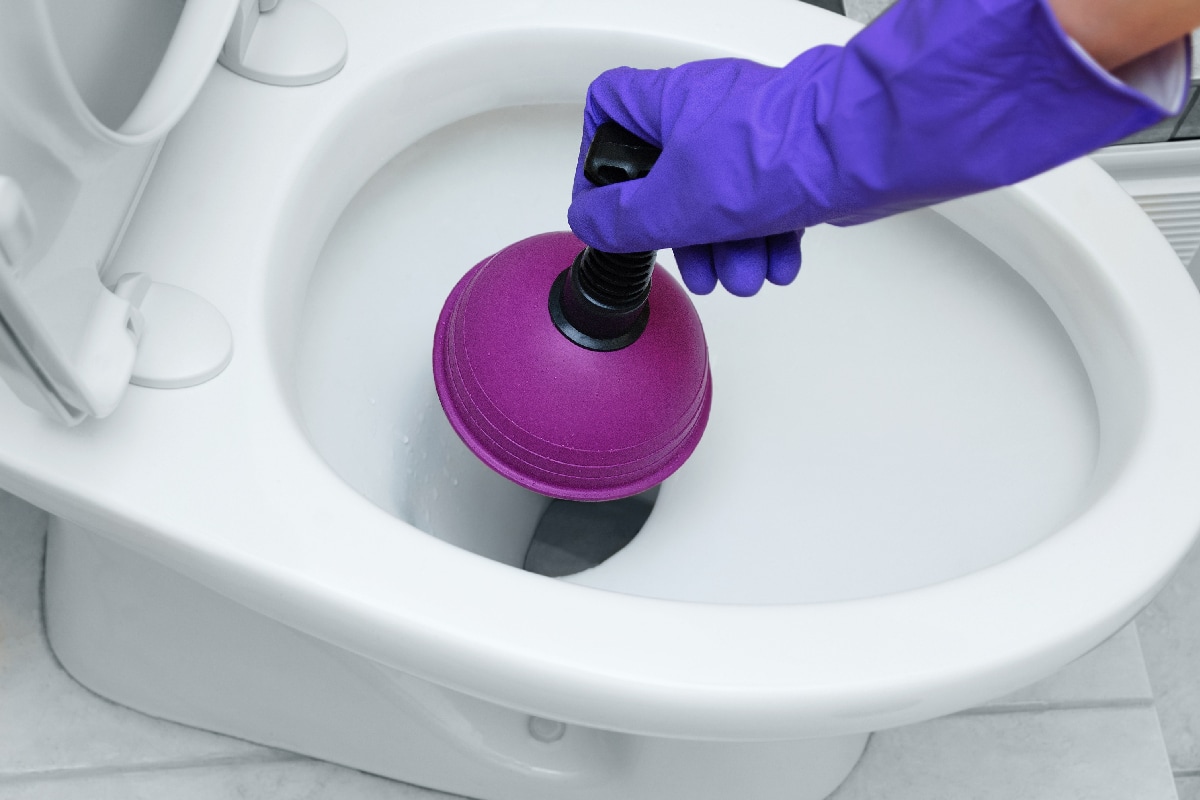 A hand in a protective glove cleans blockage in the toilet bowl with a plunger