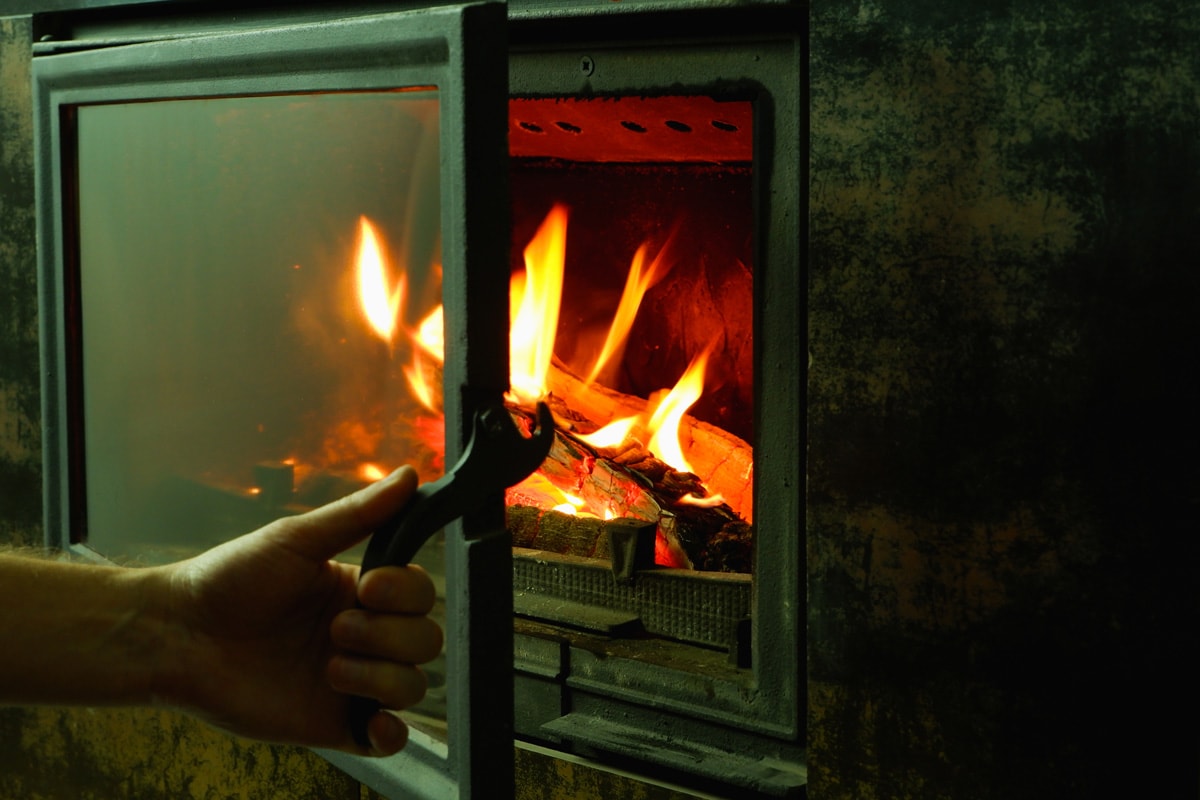 A man's hand opens the glass door of the fireplace in which the wood is burning.