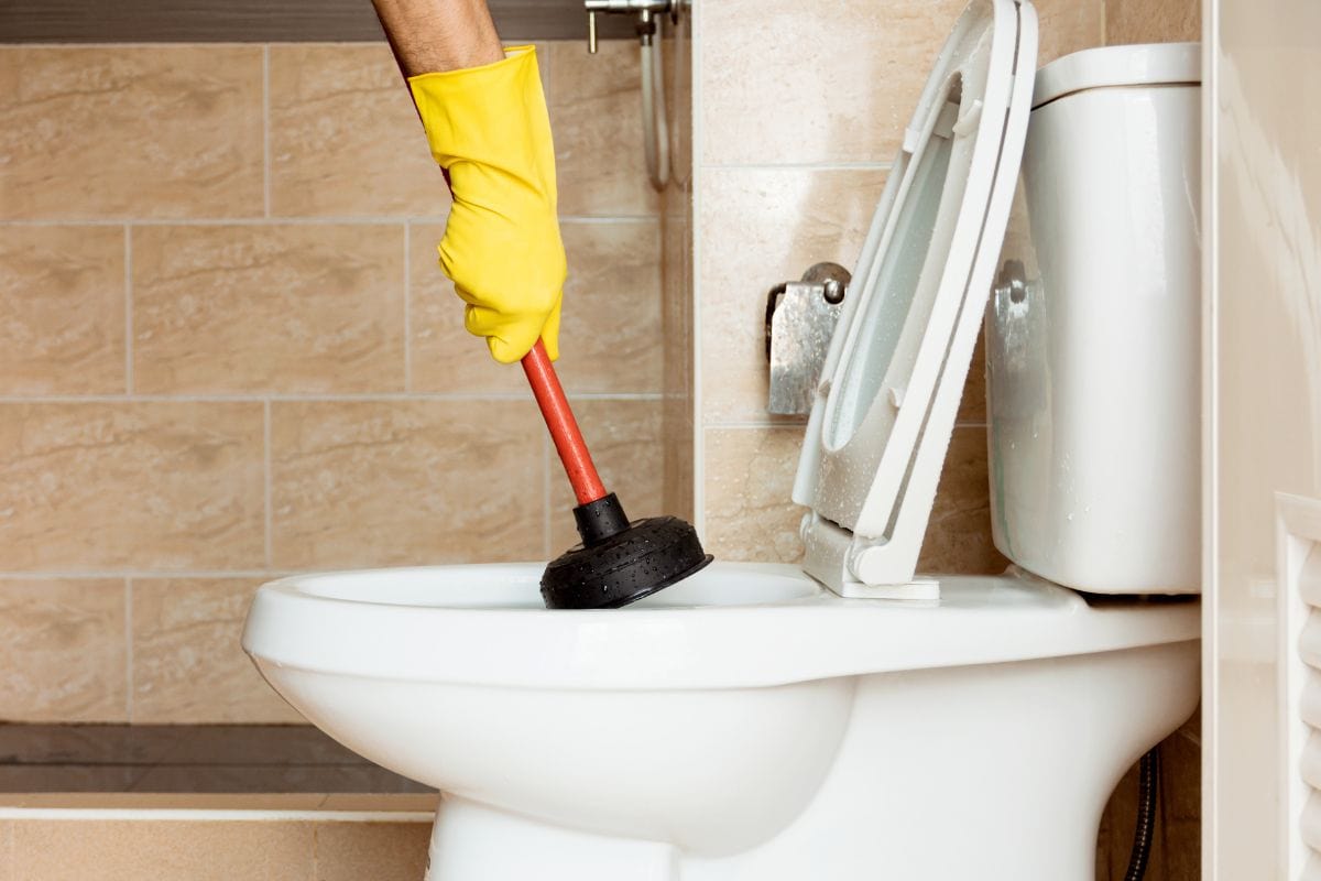 Human hand wearing yellow rubber gloves is using a device to fix a clogged toilet bowl.