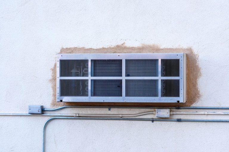 New PTAC packaged terminal air conditioner unit covered with rear grille installed through a commercial building exterior wall. Electrical conduit pipes and boxes on unpainted wall surface. PTAC Unit Smells Musty - Why And What To Do?