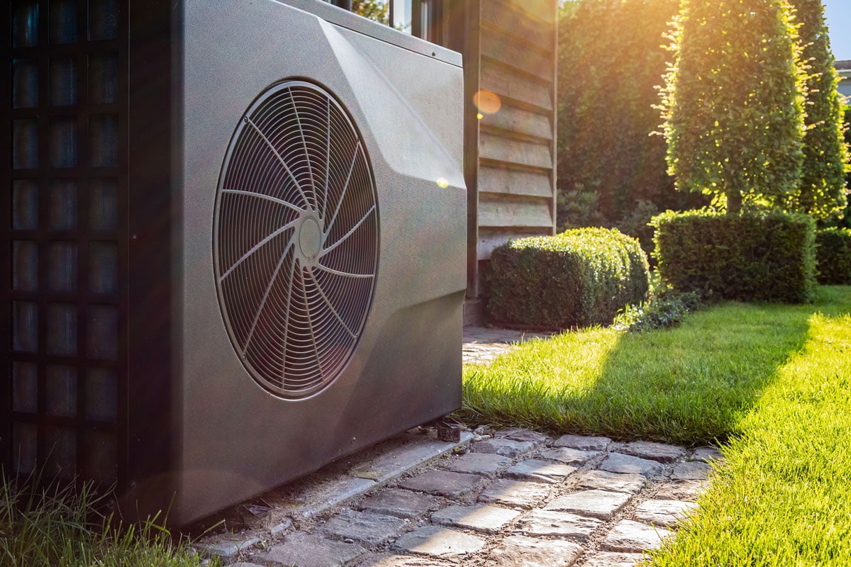 black full inverter heat pump outside in the garden, near wooden pool house on a sunny day