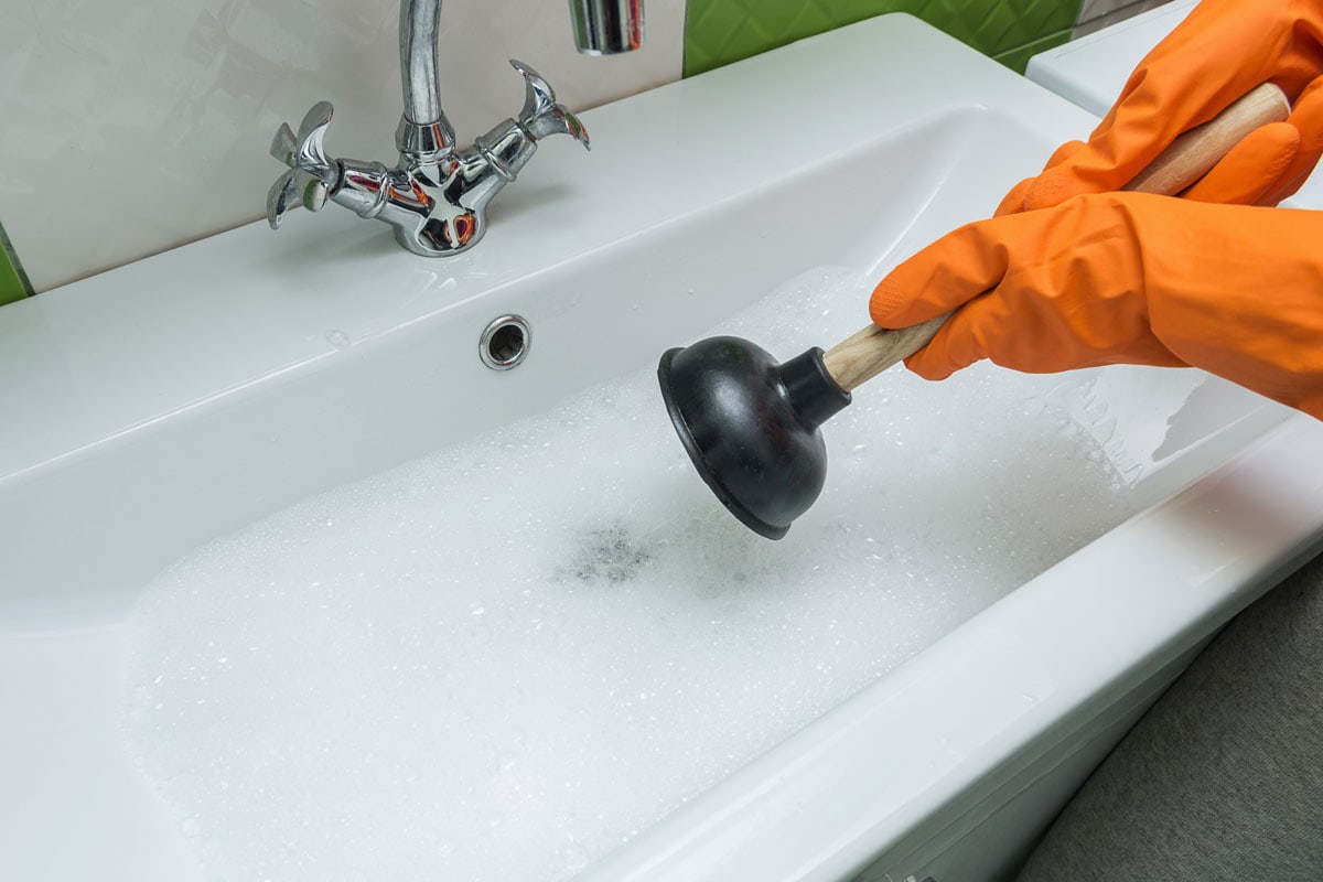 photo of a man wearing orange rubber safety gloves plunger a kitchen sink