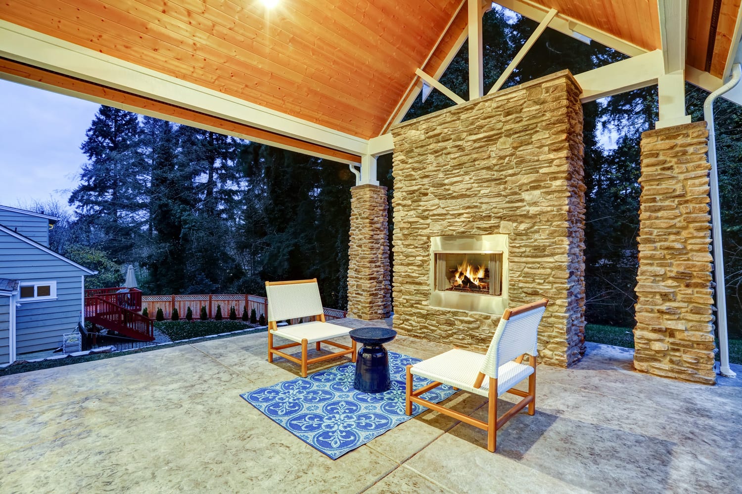 Chic covered back patio with built in gas fireplace, stone pillars, plank vaulted ceiling over cozy teak wood sofa set topped with white cushions and green pillows.