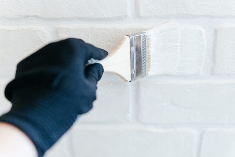 Close-up of a worker's hand with a brush painting a brick wall with white paint. How To Apply Drylok [A Detailed Guide For Beginners]