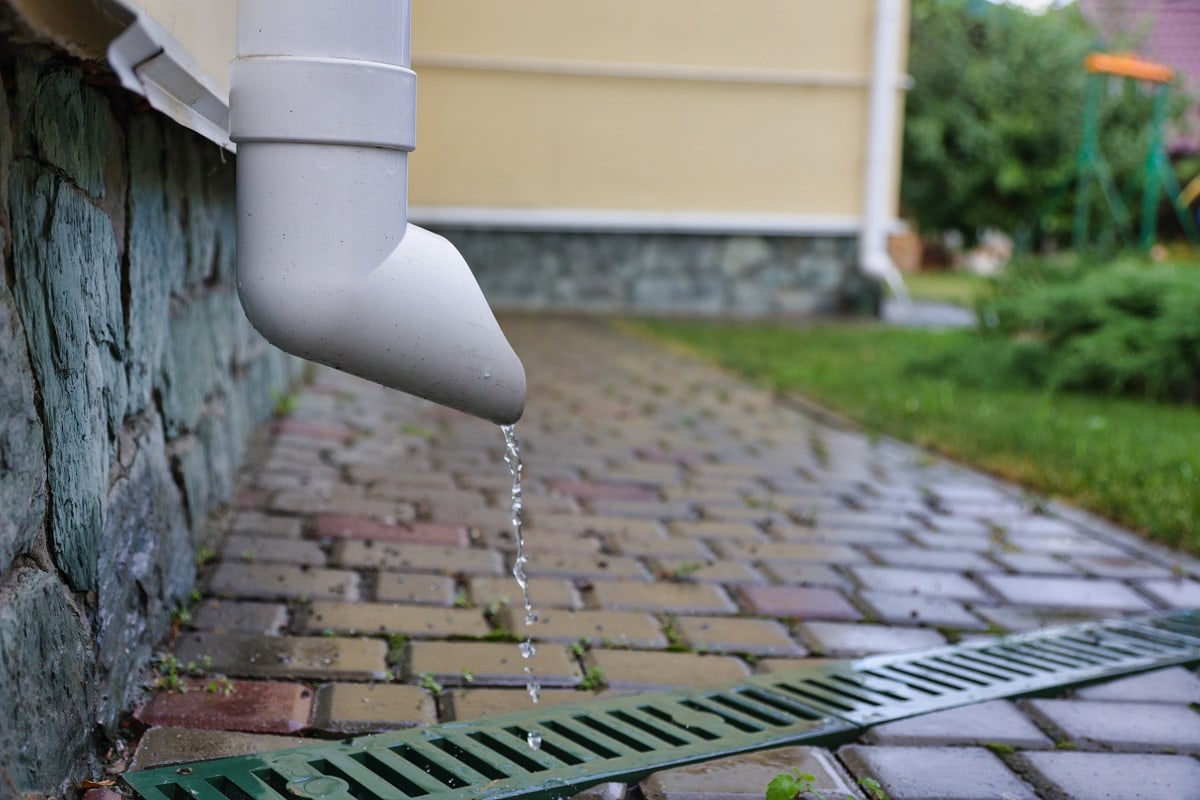 How Far Should The AC Drain Line Be From The House - drainpipe of a beige house into a gutter with a grate,