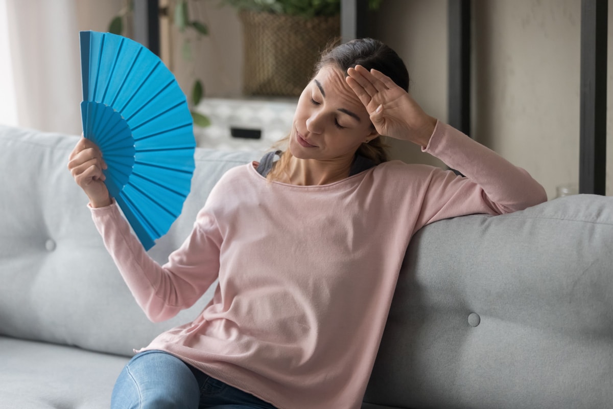 Overheated female sitting on couch in living room at hot summer weather day feeling discomfort suffers from heat waving blue fan to cool herself