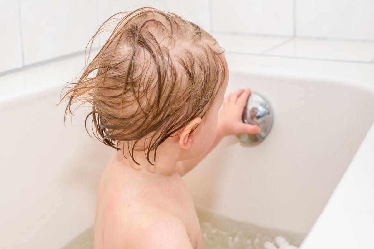 Wet baby girl with blond hair looks at her reflection in overflow drain knob of the bathtub